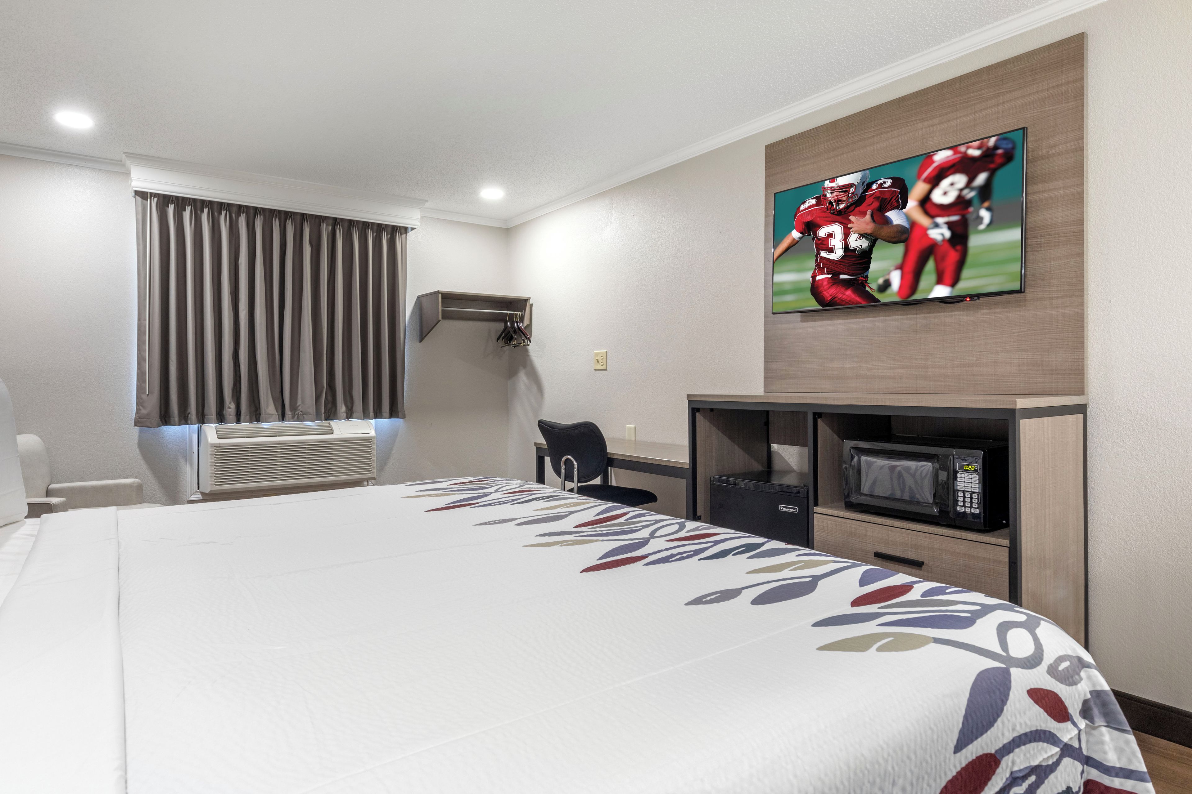 Habitación de hotel con cama, televisor de pantalla plana montado en la pared, escritorio con silla, microondas y aire acondicionado.