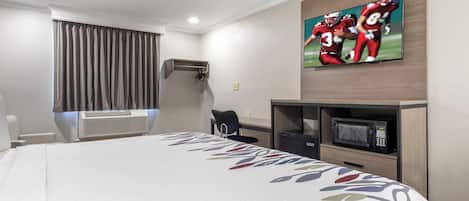 A hotel room with a bed, a flat-screen TV mounted on the wall, a desk with a chair, a microwave, and an air conditioning unit.