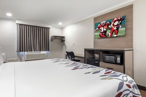Habitación de hotel con cama, televisor de pantalla plana montado en la pared, escritorio con silla, microondas y aire acondicionado.