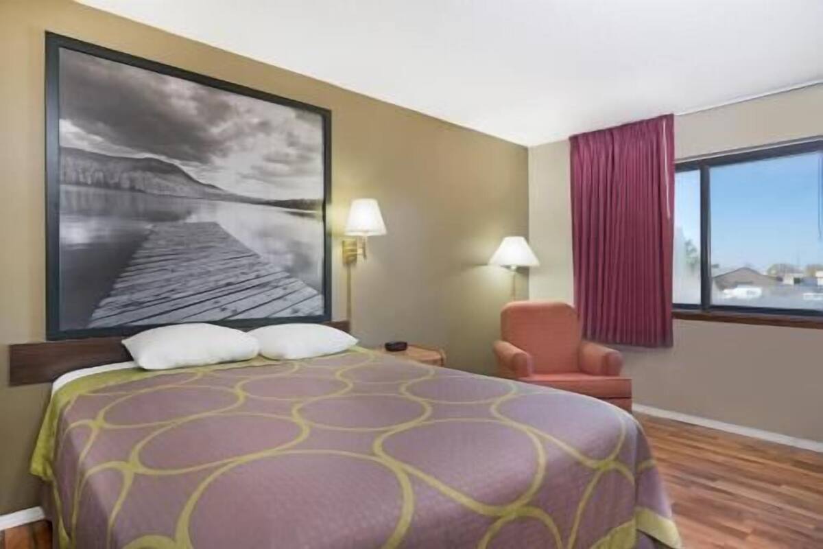 Ein Hotelzimmer mit Bett, Sessel, Nachttisch, Lampe, einem Fenster mit Vorhängen und einem großen Wandbild.