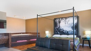 Una habitación con cama con dosel, una zona de estar con mesa, una bañera con detalles de mosaico y un ventanal con vistas a árboles.