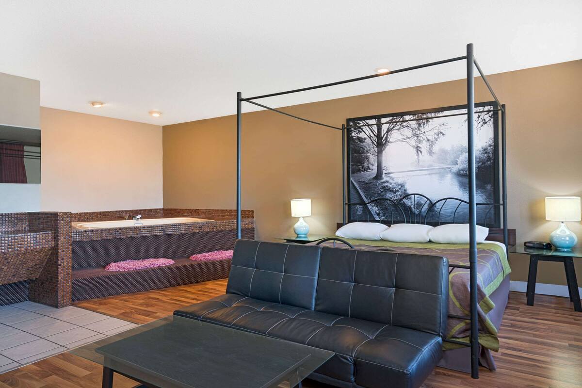 Ein Zimmer mit Himmelbett, einer Sitzecke mit Tisch, einer Badewanne mit Mosaikverkleidung und einem großen Fenster mit Blick auf Bäume.