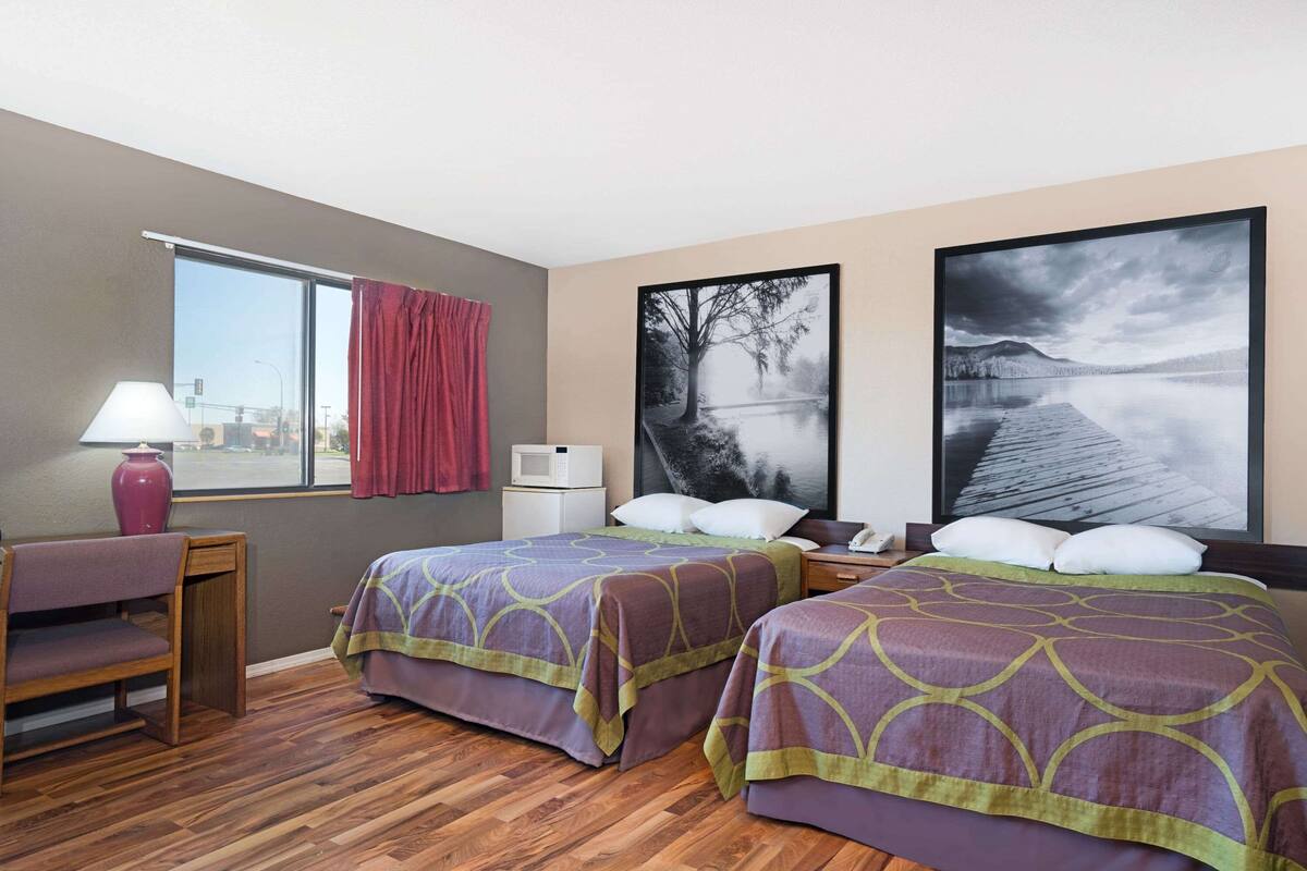 Ein Hotelzimmer mit zwei Betten, einem Schreibtisch, einem Stuhl, einem Fenster mit Vorhängen, einer Lampe, einer Mikrowelle und zwei gerahmten Bildern an der Wand.