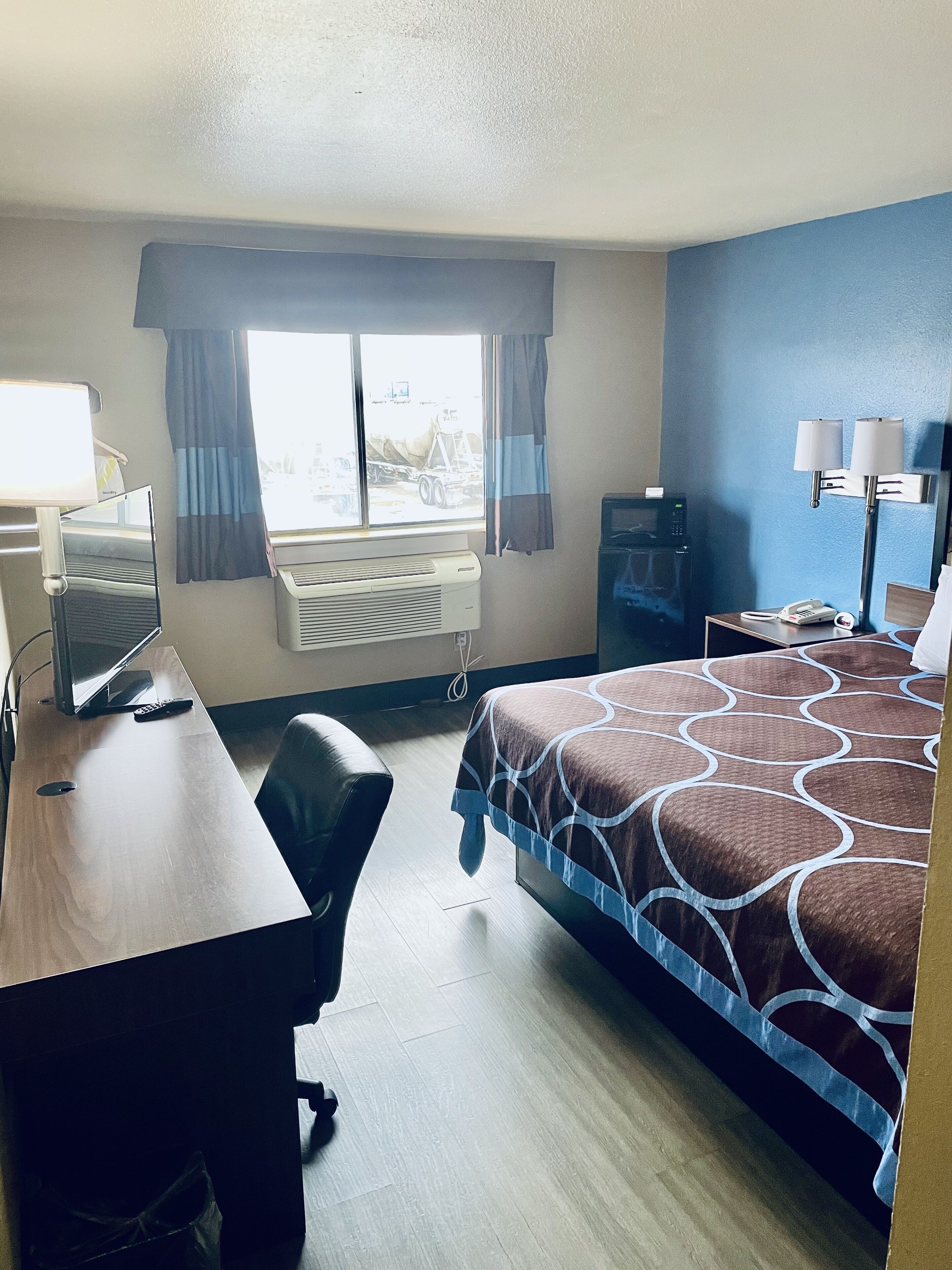 A hotel room with a bed, desk, chair, air conditioning unit, window with curtains, and a television.