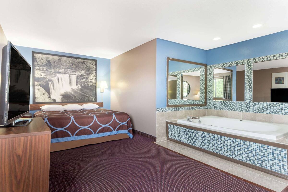 Una habitación de hotel con cama, televisor, un espejo grande y baño con bañera y paredes de azulejos.