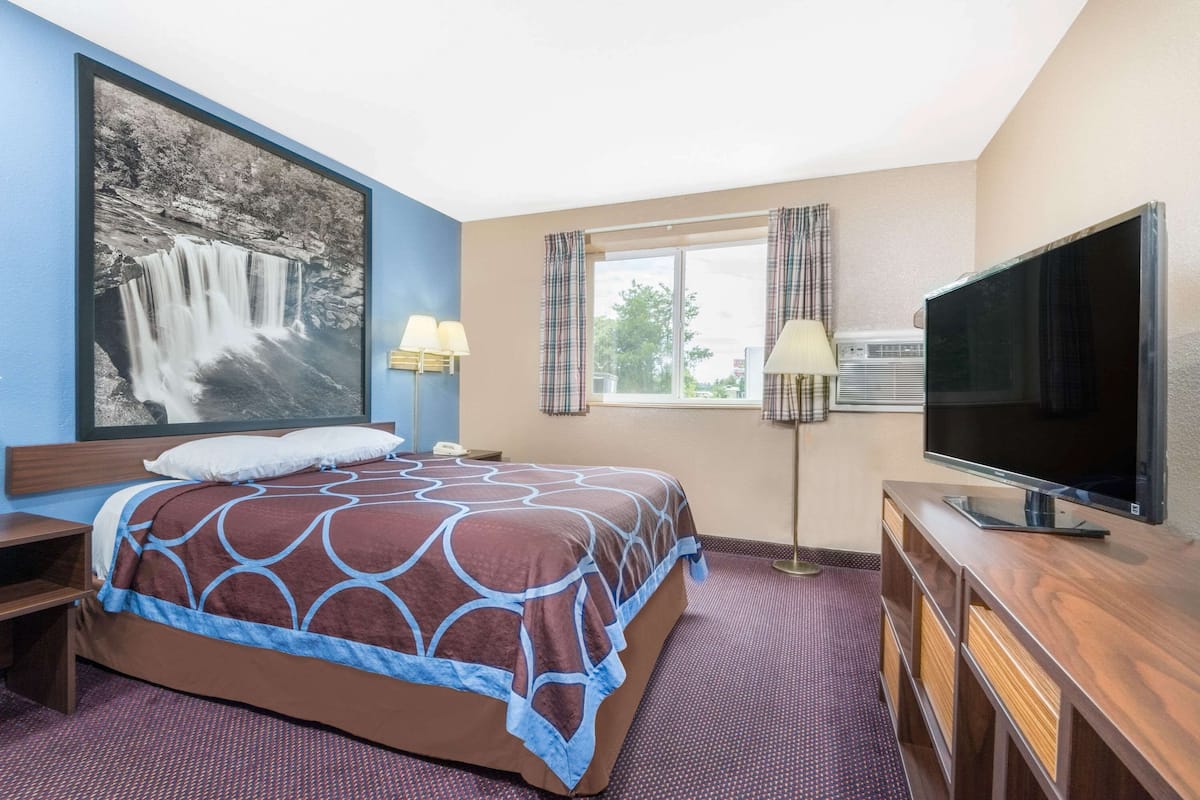 Una habitación de hotel con una cama, televisión, ventana con cortinas, un cuadro en la pared y una lámpara.