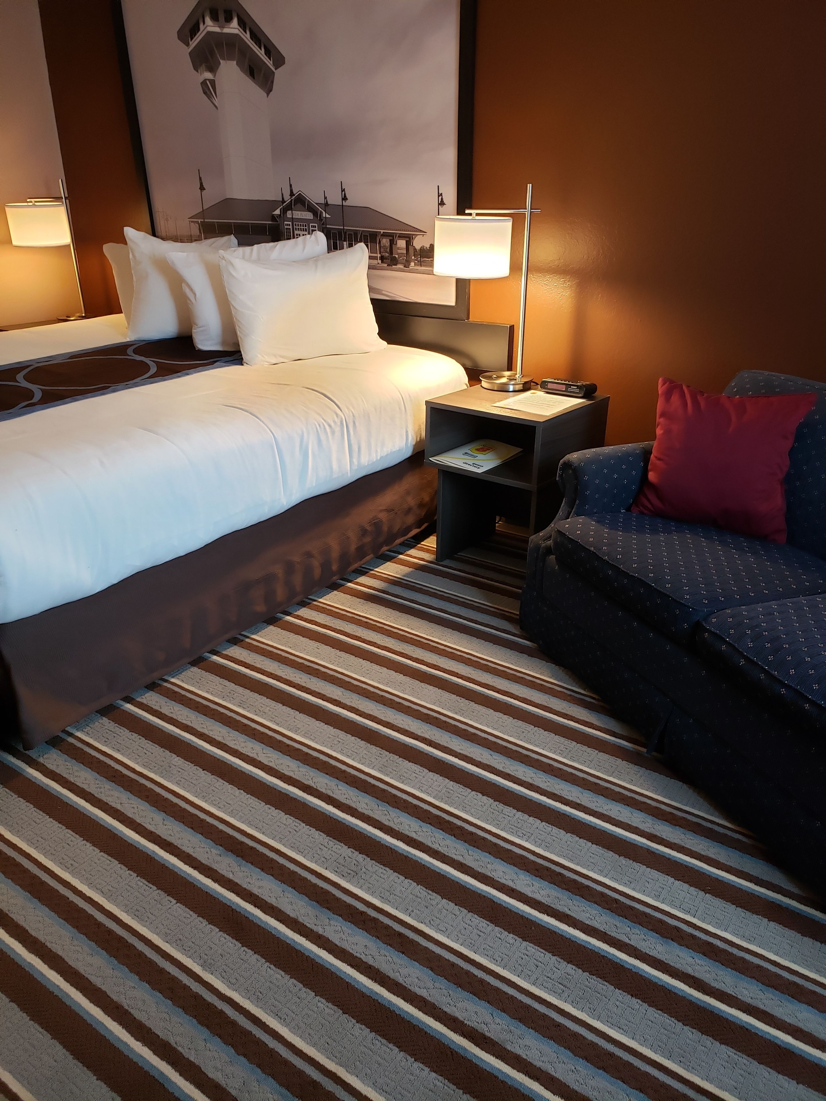 A hotel room with a large bed, a nightstand with a lamp, a sofa, and a striped carpet.