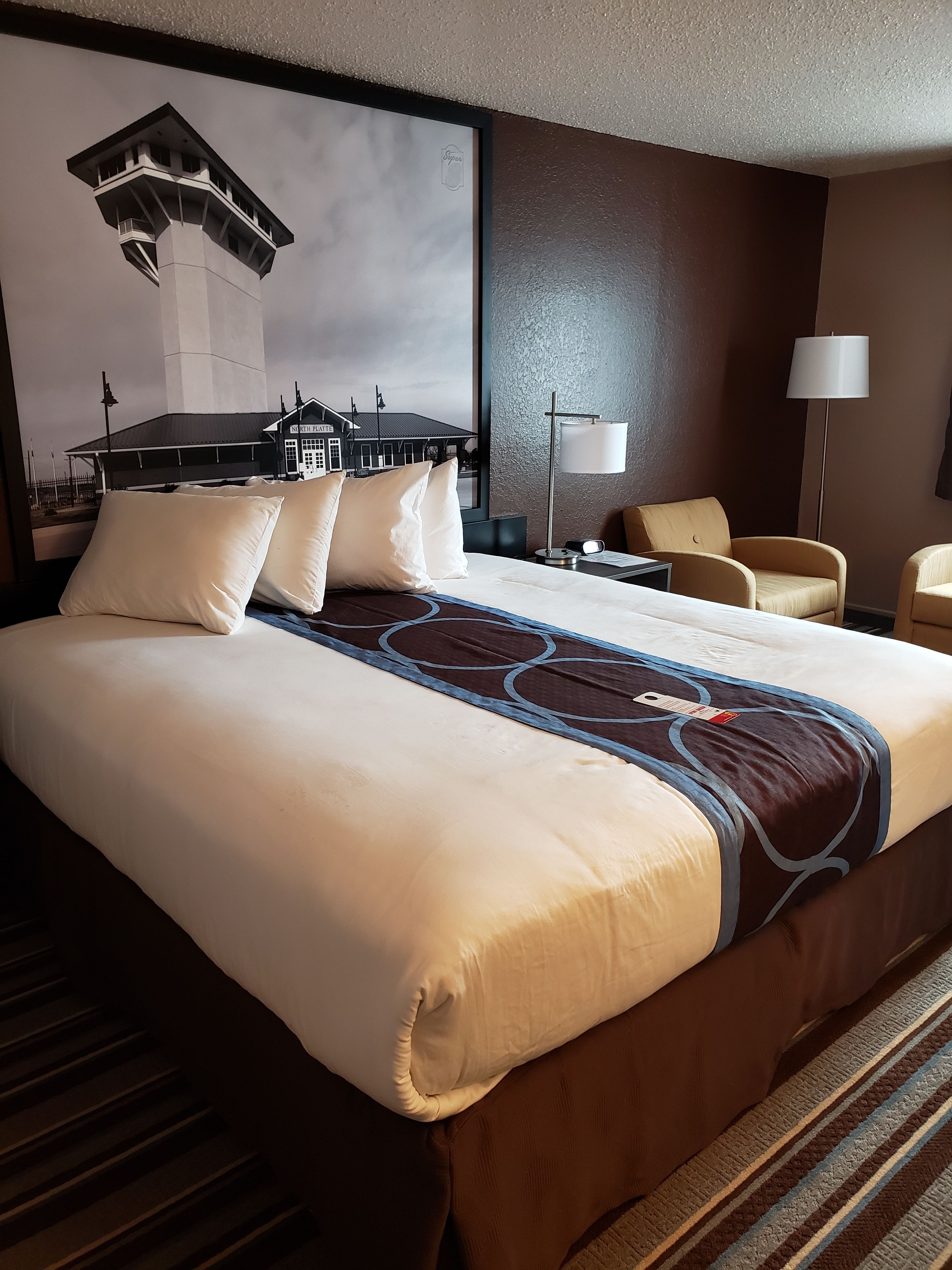 A hotel room with a large bed, two chairs, a desk, a lamp, and a wall-mounted artwork of a train station.