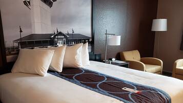 A hotel room with a large bed, two chairs, a desk, a lamp, and a wall-mounted artwork of a train station.
