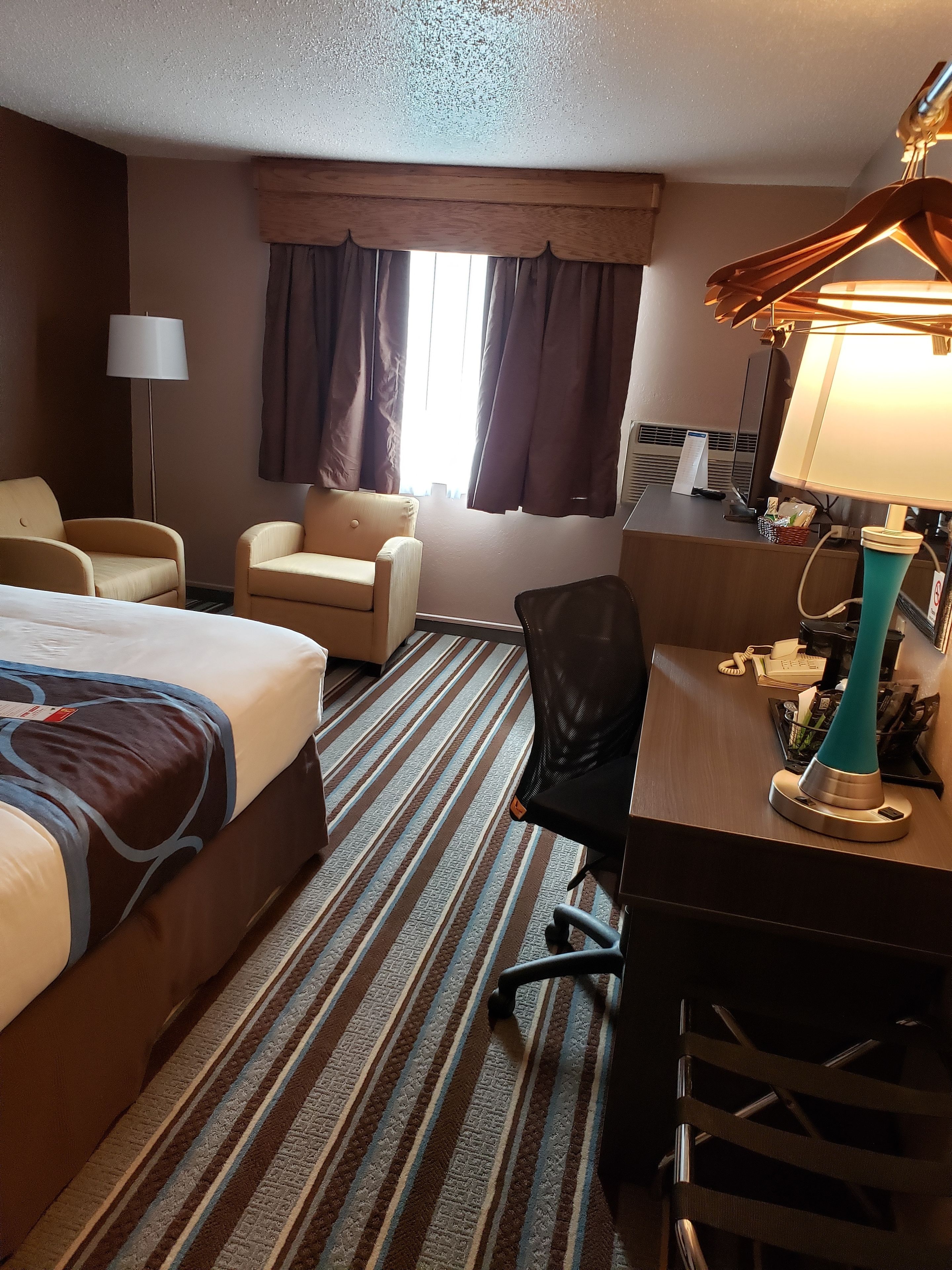 Habitación de hotel con una cama, dos sillones, un escritorio con lámpara, una computadora y un ventanal con cortinas.