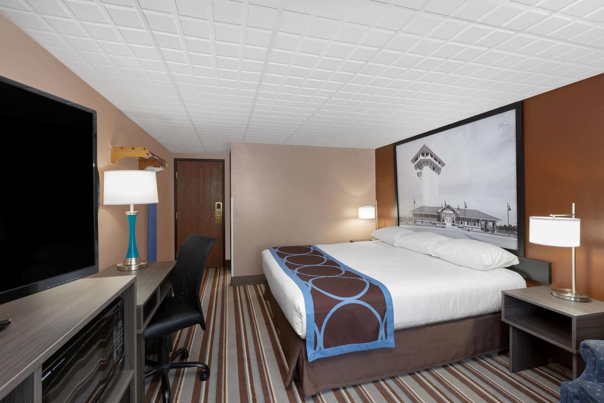 A hotel room with a bed, a desk with a flat-screen TV, a chair, a lamp, and a framed picture on the wall.