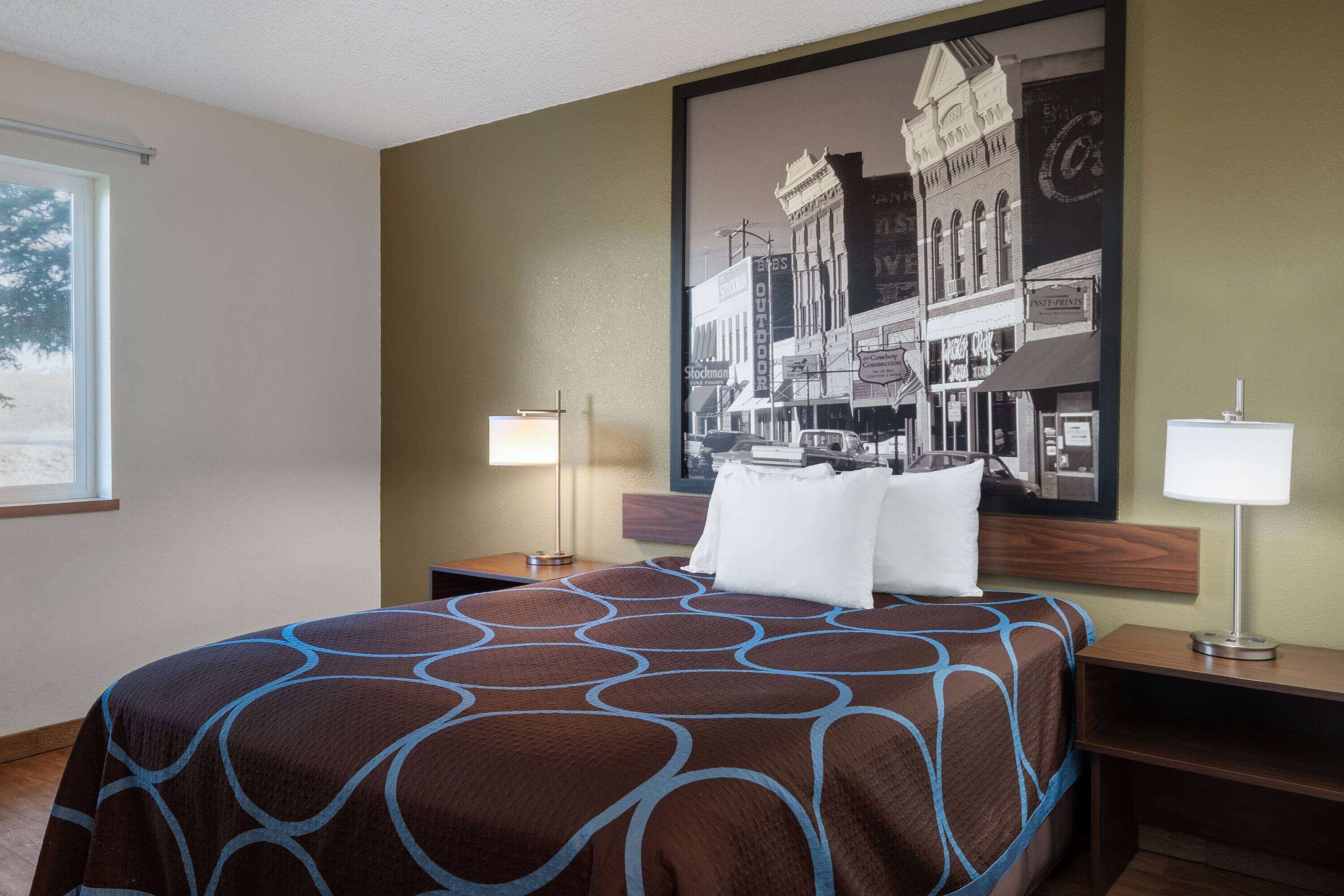 Habitación de hotel con una cama grande, dos mesitas de noche, una lámpara de noche, una ventana y un cuadro colgado en la pared con una escena callejera.