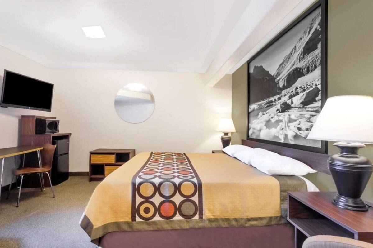 A hotel room with a bed, a desk, a chair, a television, a mirror, a lamp, and a framed picture on the wall.