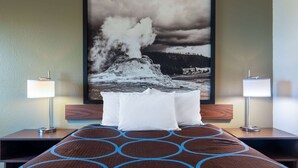 A hotel room with a bed, two bedside lamps, a large framed picture of a geyser, and a patterned rug. - Super 8 by Wyndham Livingston Yellowstone (Livingston)
