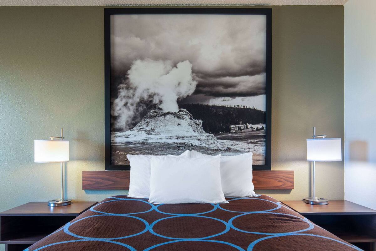 A hotel room with a bed, two bedside lamps, a large framed picture of a geyser, and a patterned rug.