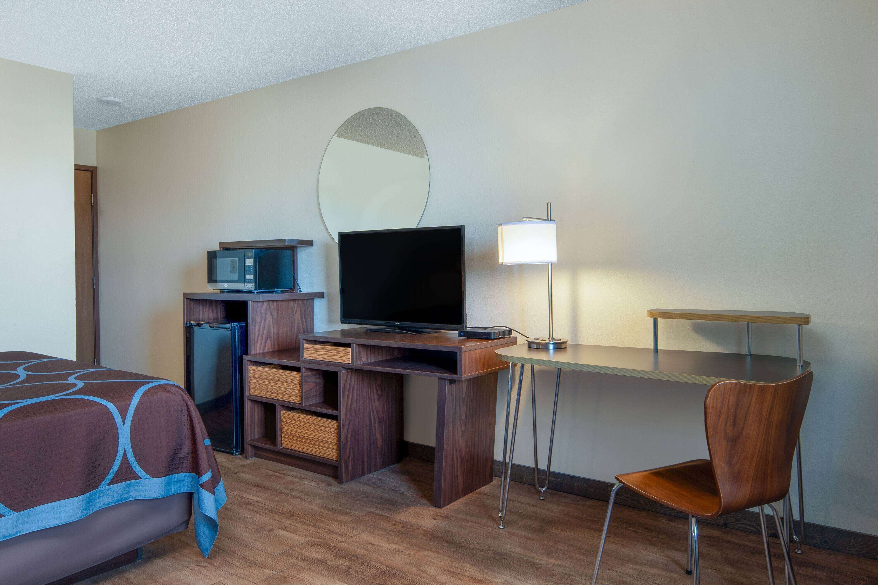 A hotel room with a bed, a desk with a chair, a television, a mirror, a lamp, and a minibar.