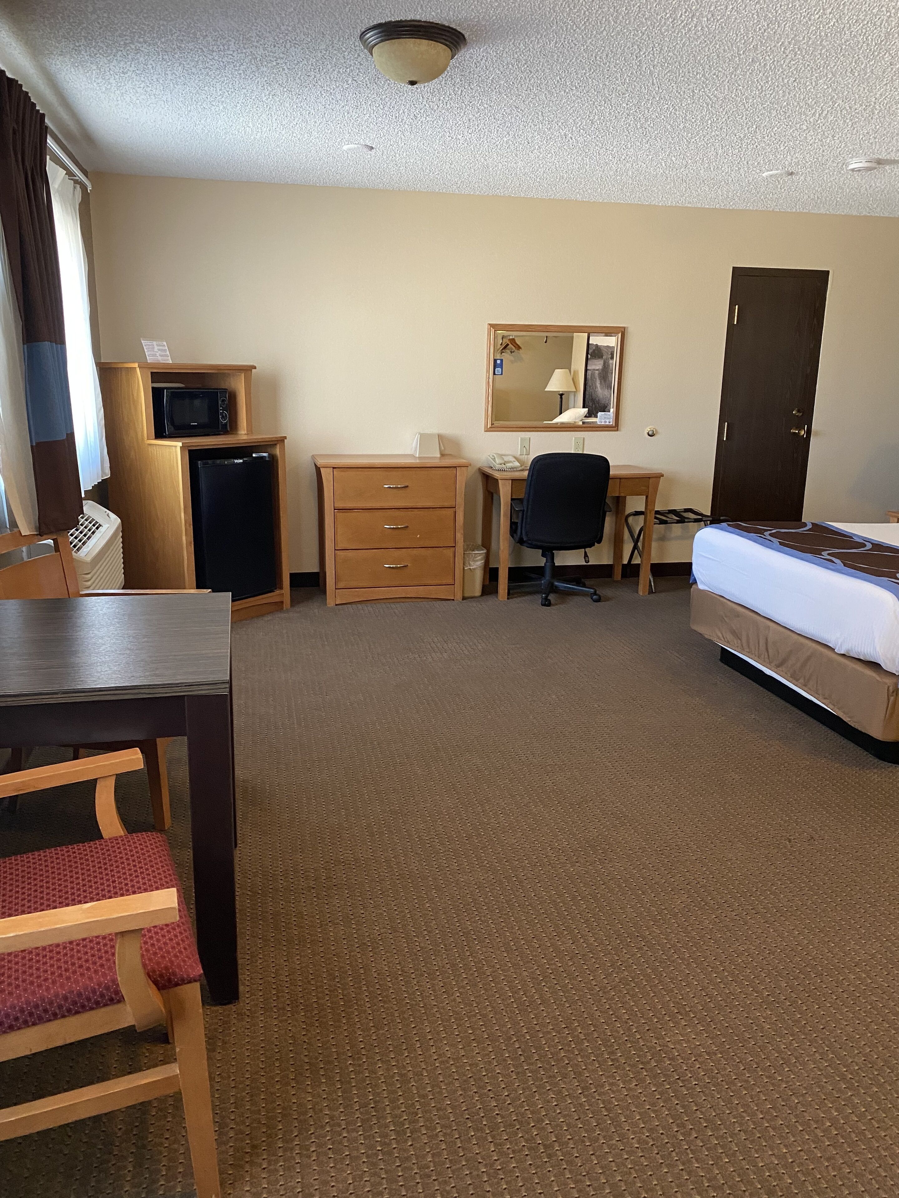 A hotel room with a bed, desk, chair, dresser, microwave, refrigerator, and a door.