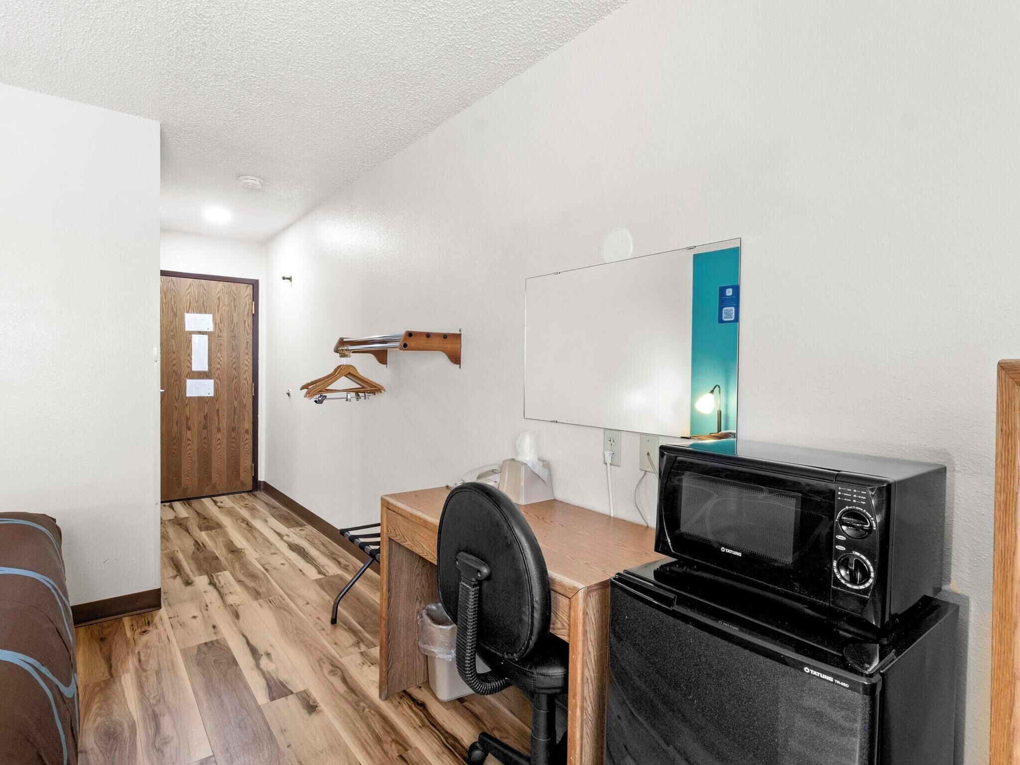 Una habitación compacta con suelo de madera, un escritorio con silla, un microondas negro, una pizarra y un perchero de madera.