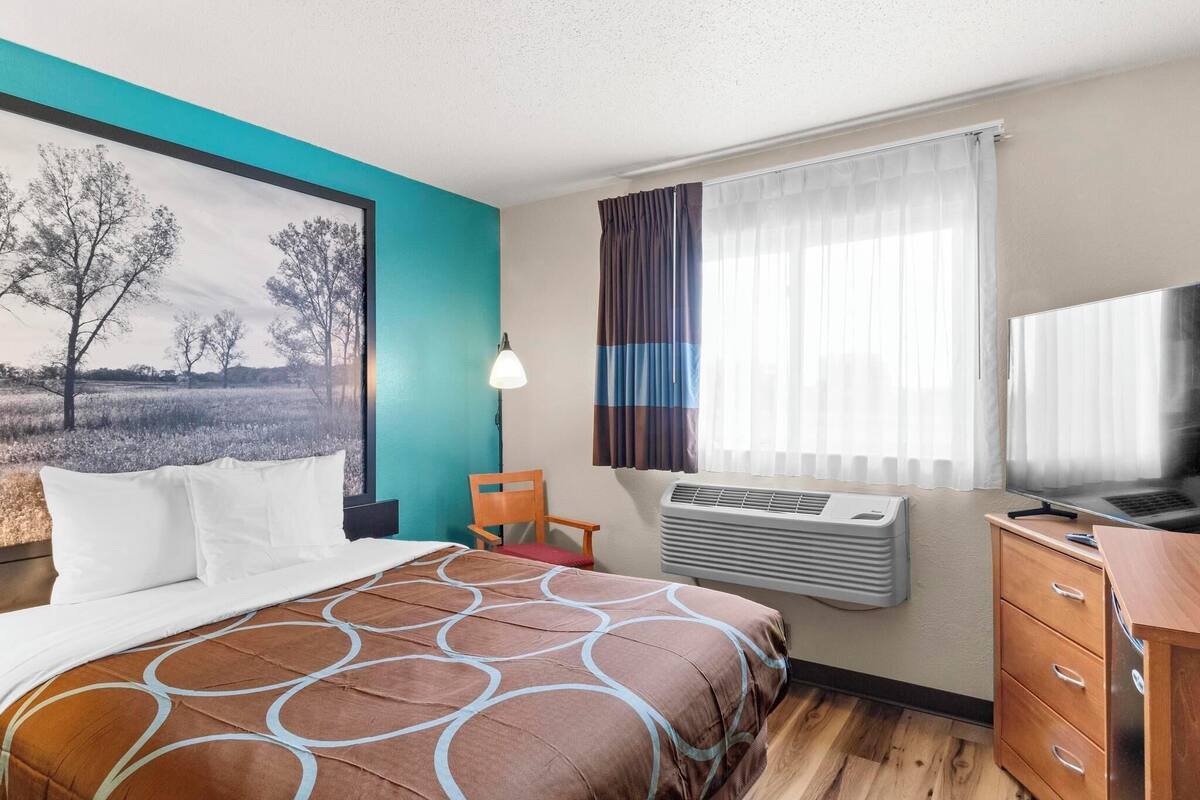 A hotel room with a bed, a desk, a chair, an air conditioner, and a TV.