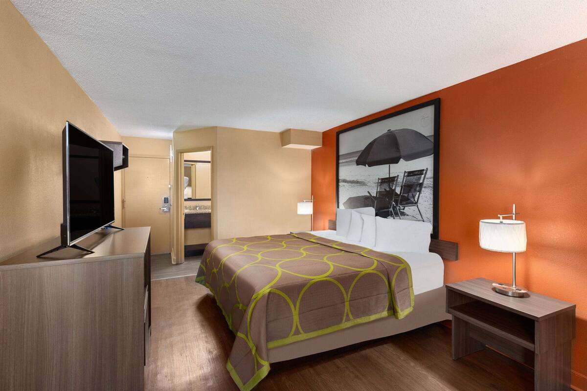 A hotel room with a bed, a TV, a desk, and a painting on the wall.