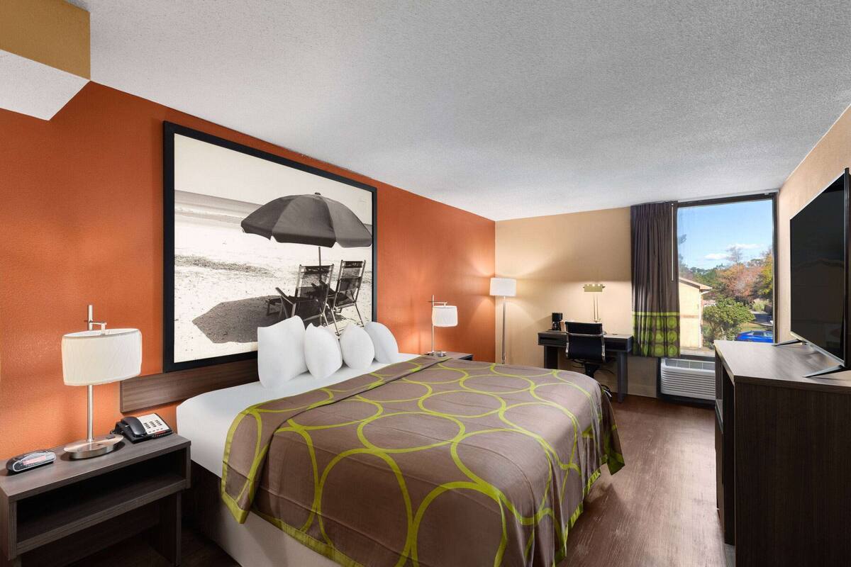 A hotel room with a bed, a desk, a chair, a TV, a window with a view, and a painting on the wall.