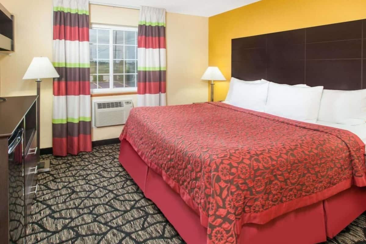 A hotel room with a bed, two bedside lamps, a TV, a window with curtains, and a carpeted floor.
