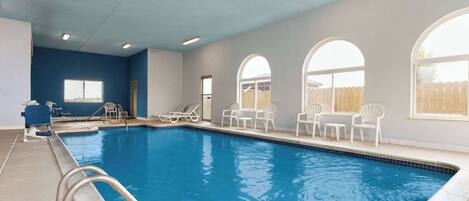 An indoor swimming pool with clear blue water, surrounded by white chairs and tables, and large arched windows.