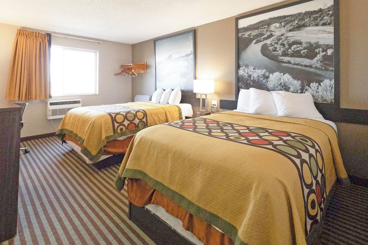 A hotel room with two beds, a large window with curtains, a wall-mounted lamp, and a framed landscape picture.