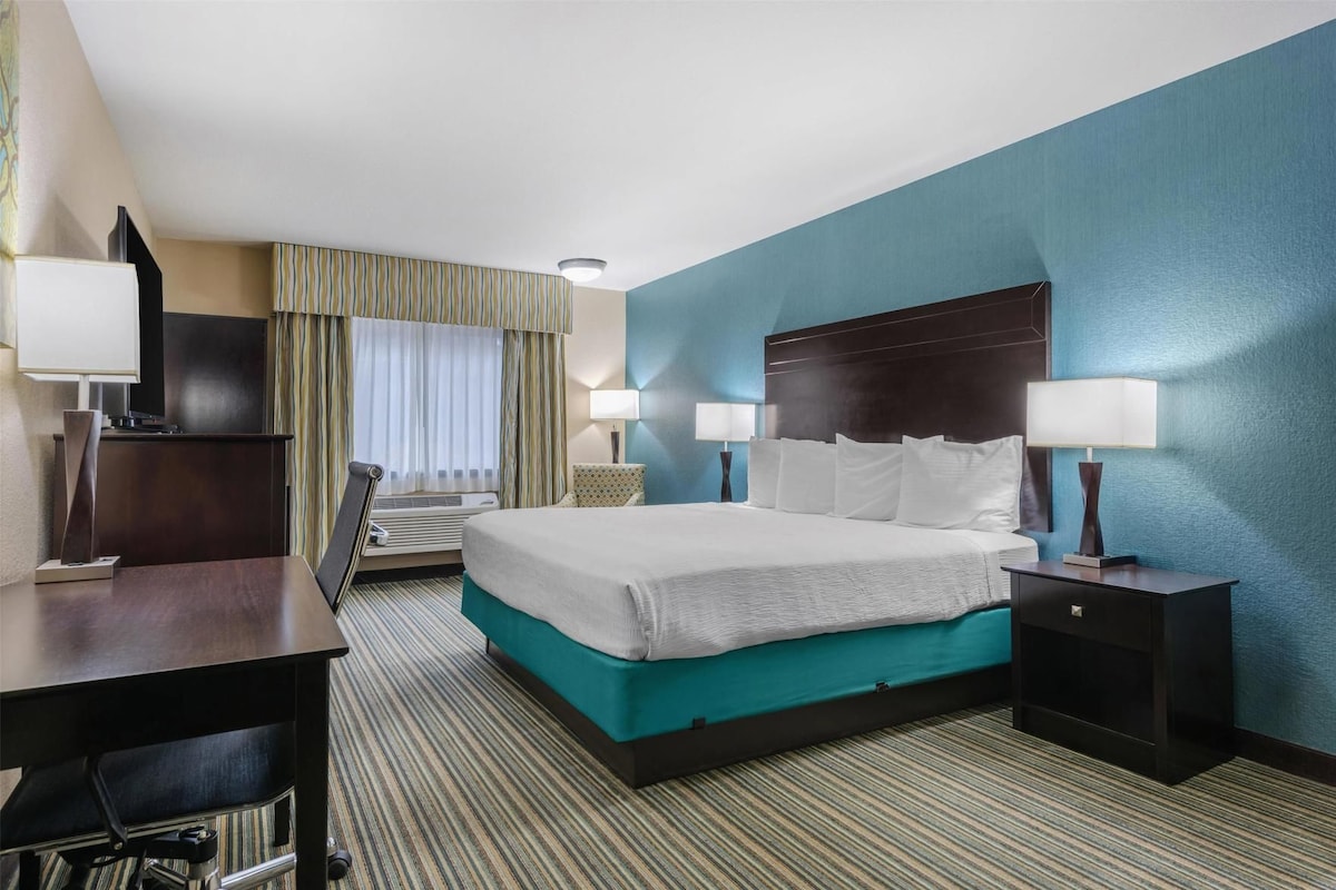 A hotel room with a bed, two bedside tables, a desk, a chair, a window with curtains, and a wall unit.