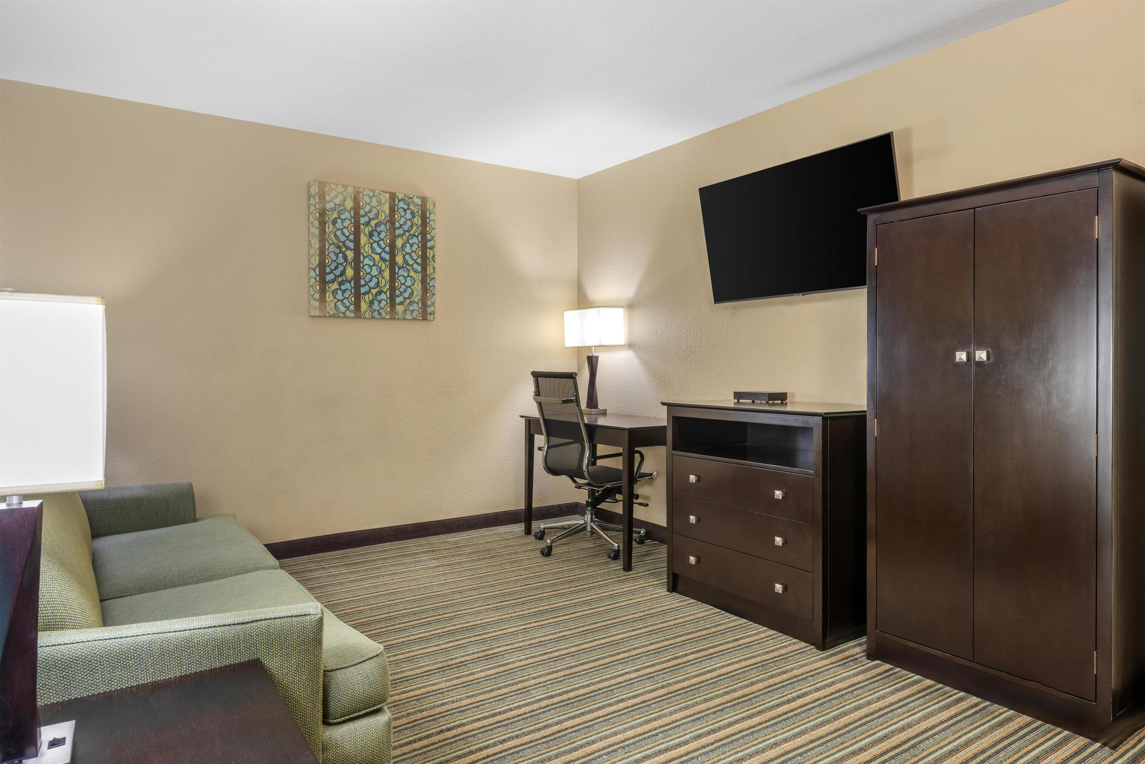 A hotel room with a green sofa, a desk with a lamp, a television on the wall, and a wardrobe.