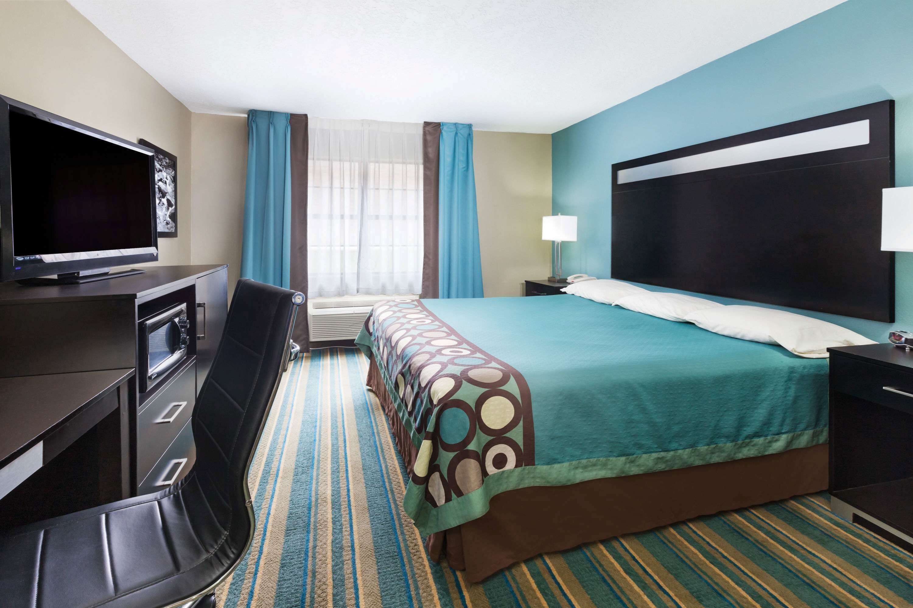 A hotel room with a bed, a desk with a TV, a chair, a lamp, and a window with curtains.