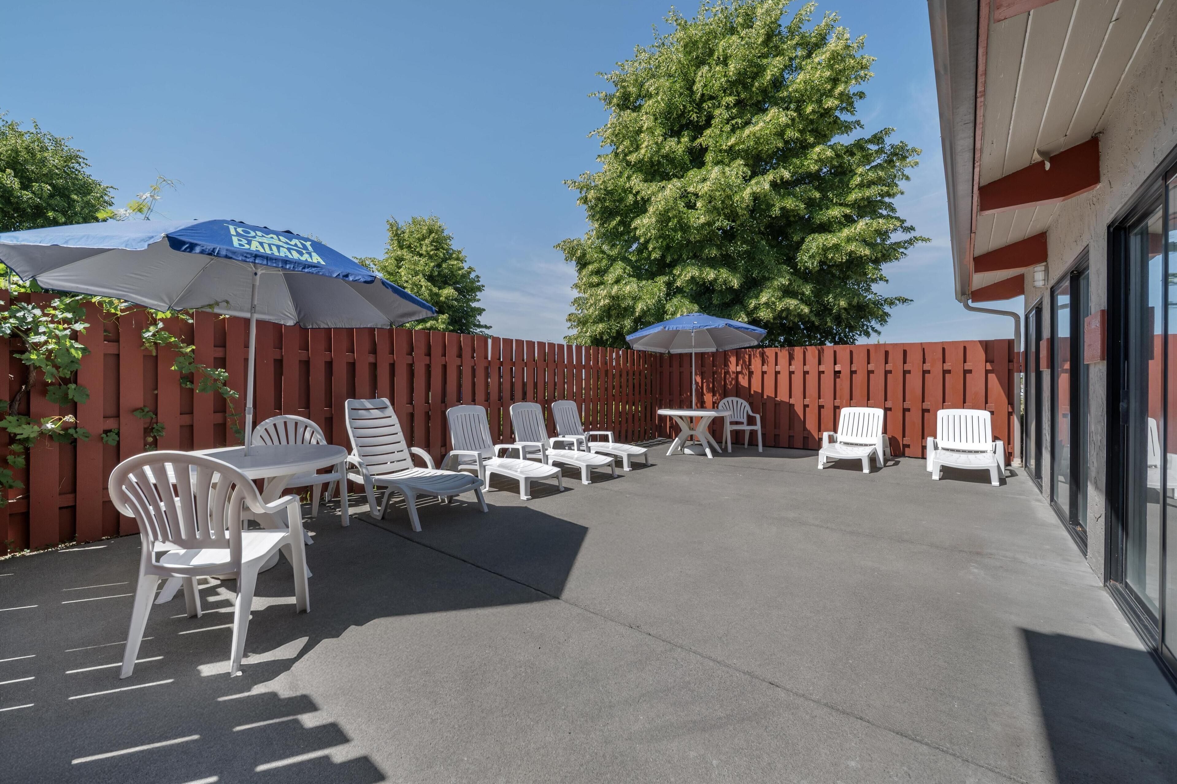 An outdoor patio with white lounge chairs and tables, blue umbrellas, and a red wooden fence.