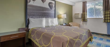 A hotel room with a large bed, a nightstand, a desk, a chair, a window with curtains, and a wall-mounted air conditioner.