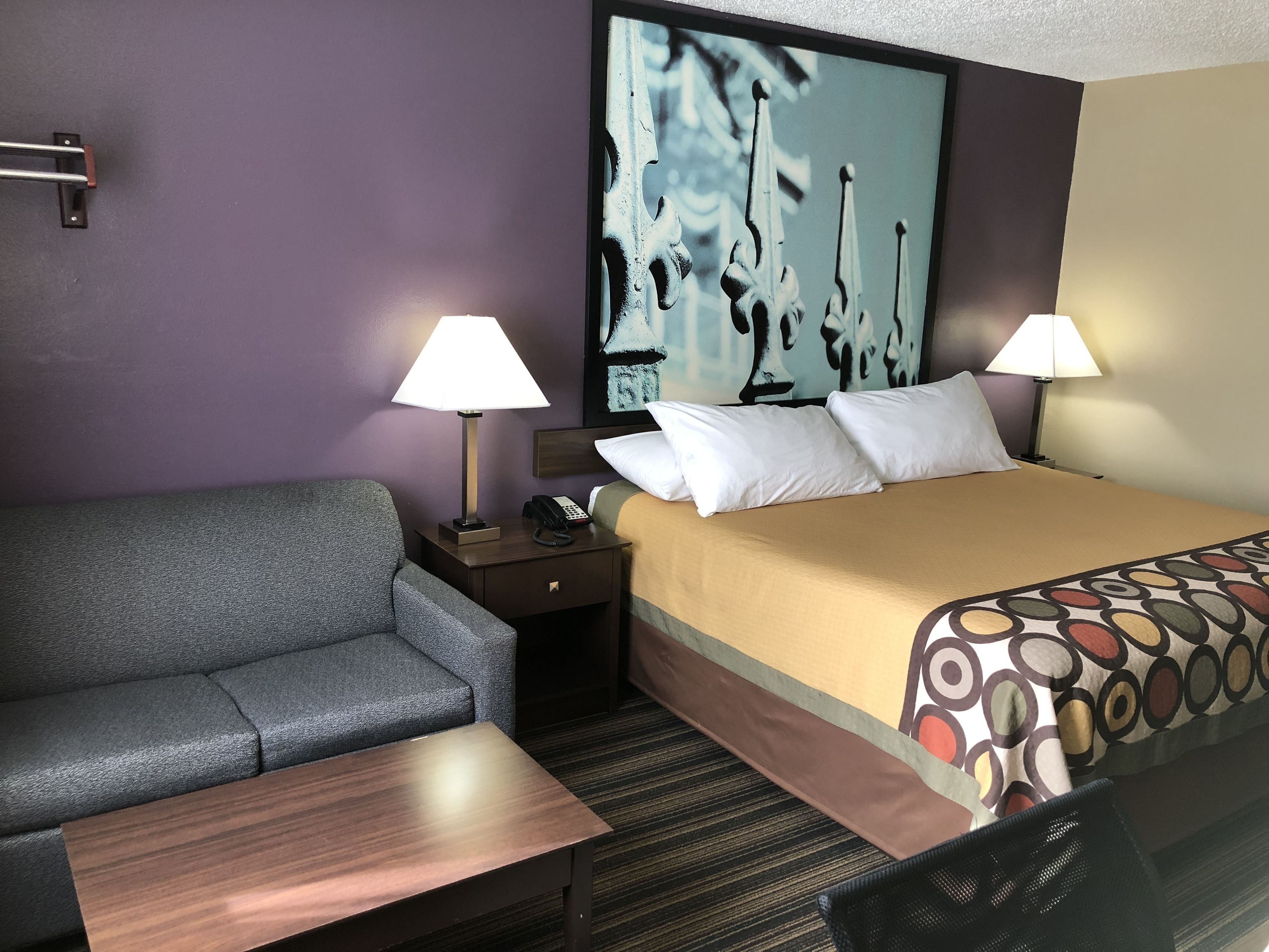 Ein Hotelzimmer mit einem Bett, zwei Nachttischlampen, einem grauen Sofa, einem Holztisch und einem großen abstrakten Gemälde an der Wand.