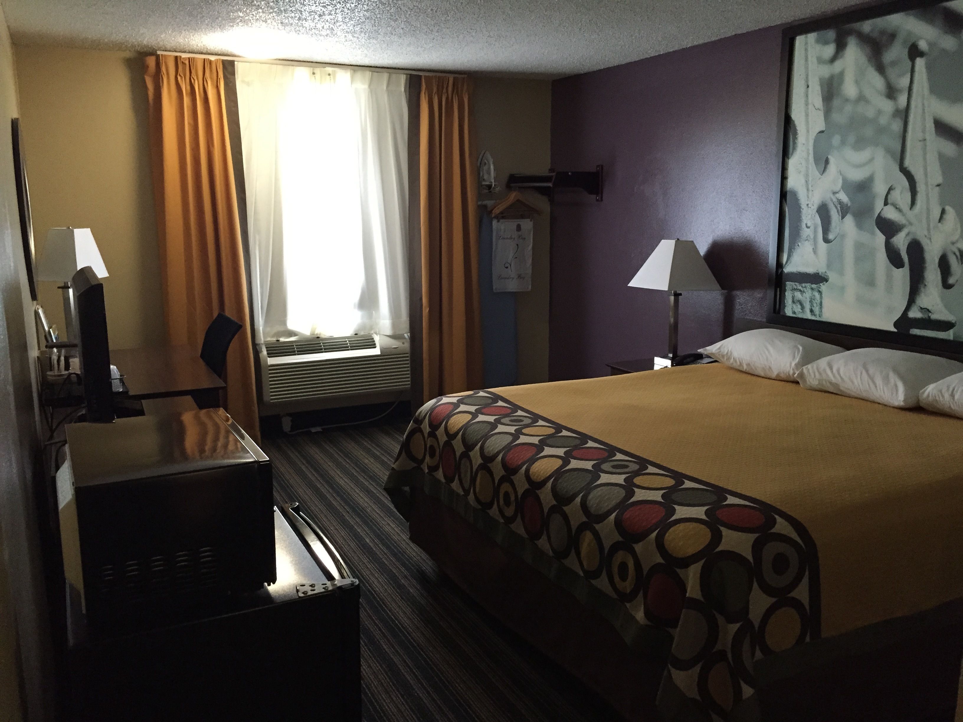 A hotel room with a bed, a desk, a chair, an air conditioner, and a painting on the wall.