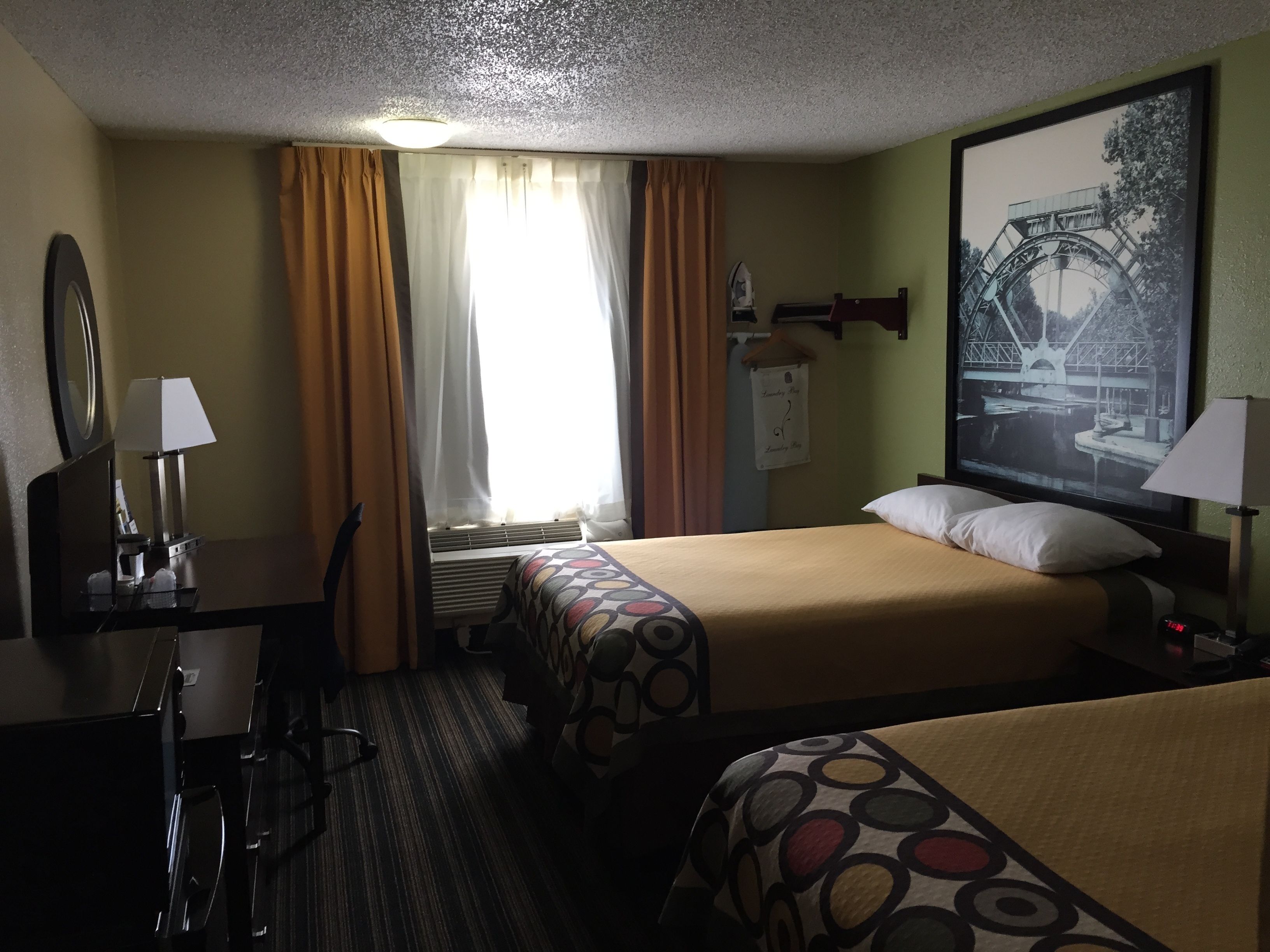 A hotel room with a bed, a desk, a chair, a mirror, a window with curtains, a wall-mounted lamp, and a framed picture.