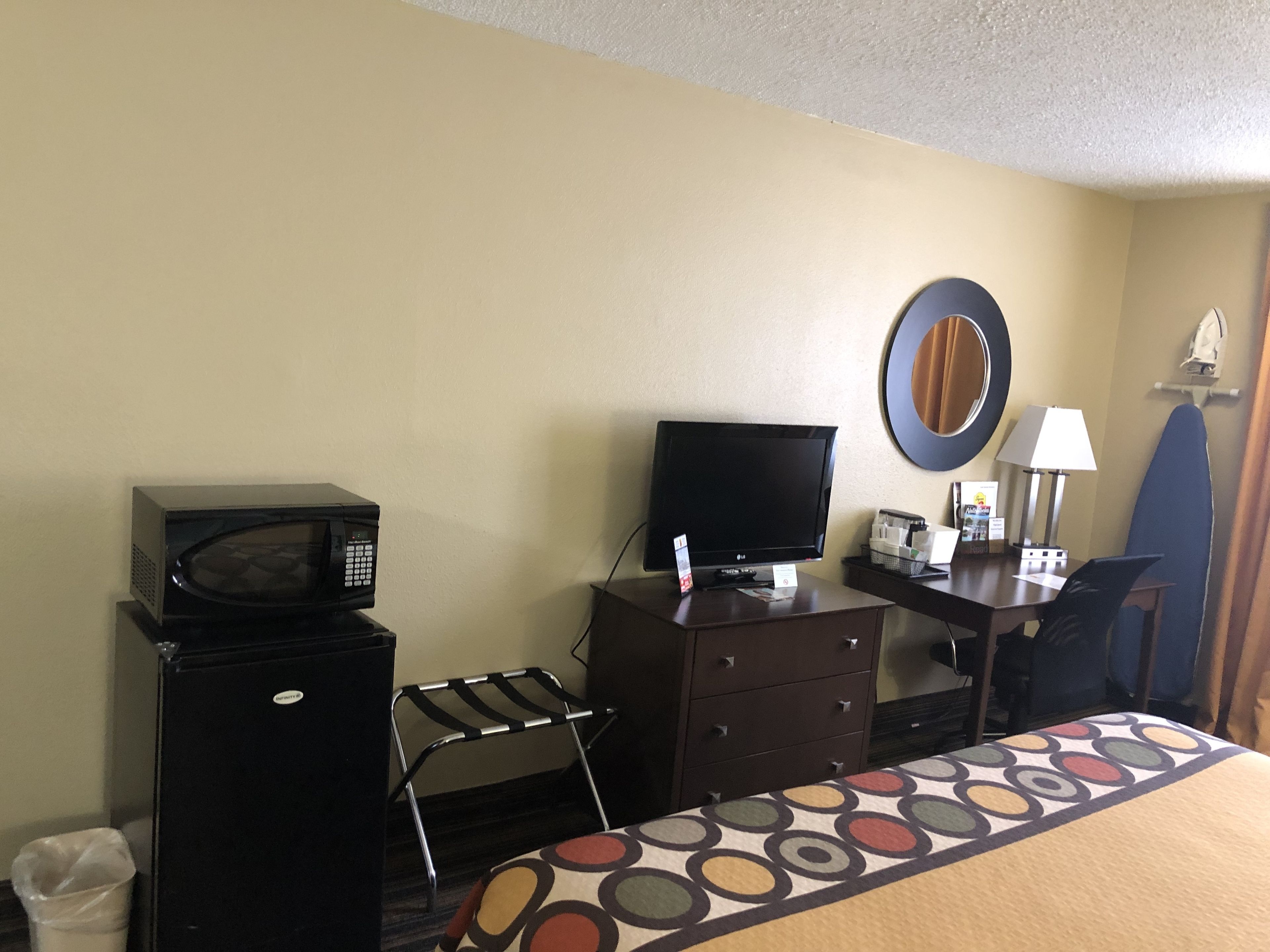 A hotel room with a bed, a desk with a television, a microwave, a chair, and a small trash bin.