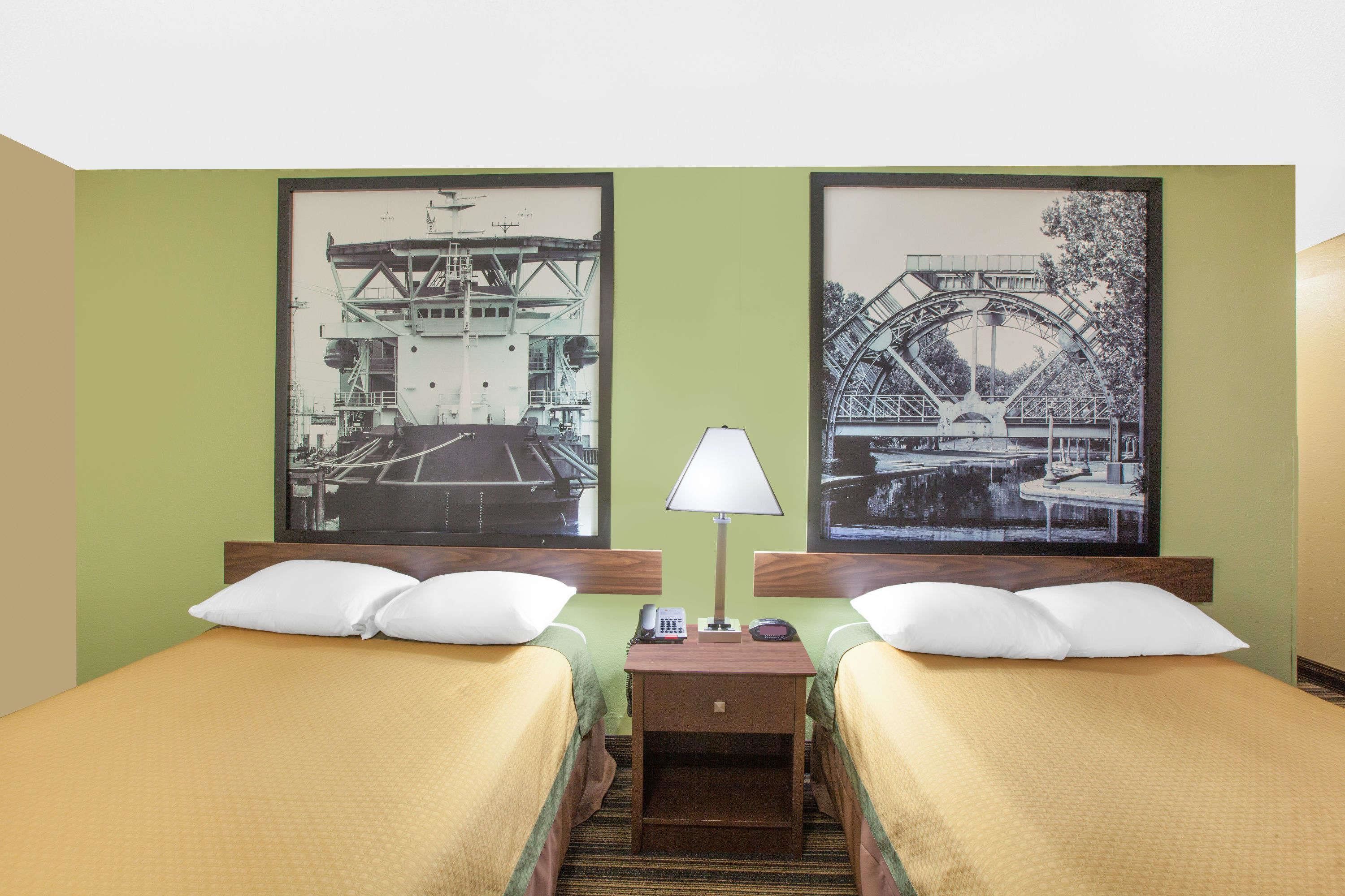 A hotel room with two beds, a nightstand with a lamp, and two framed pictures of bridges.