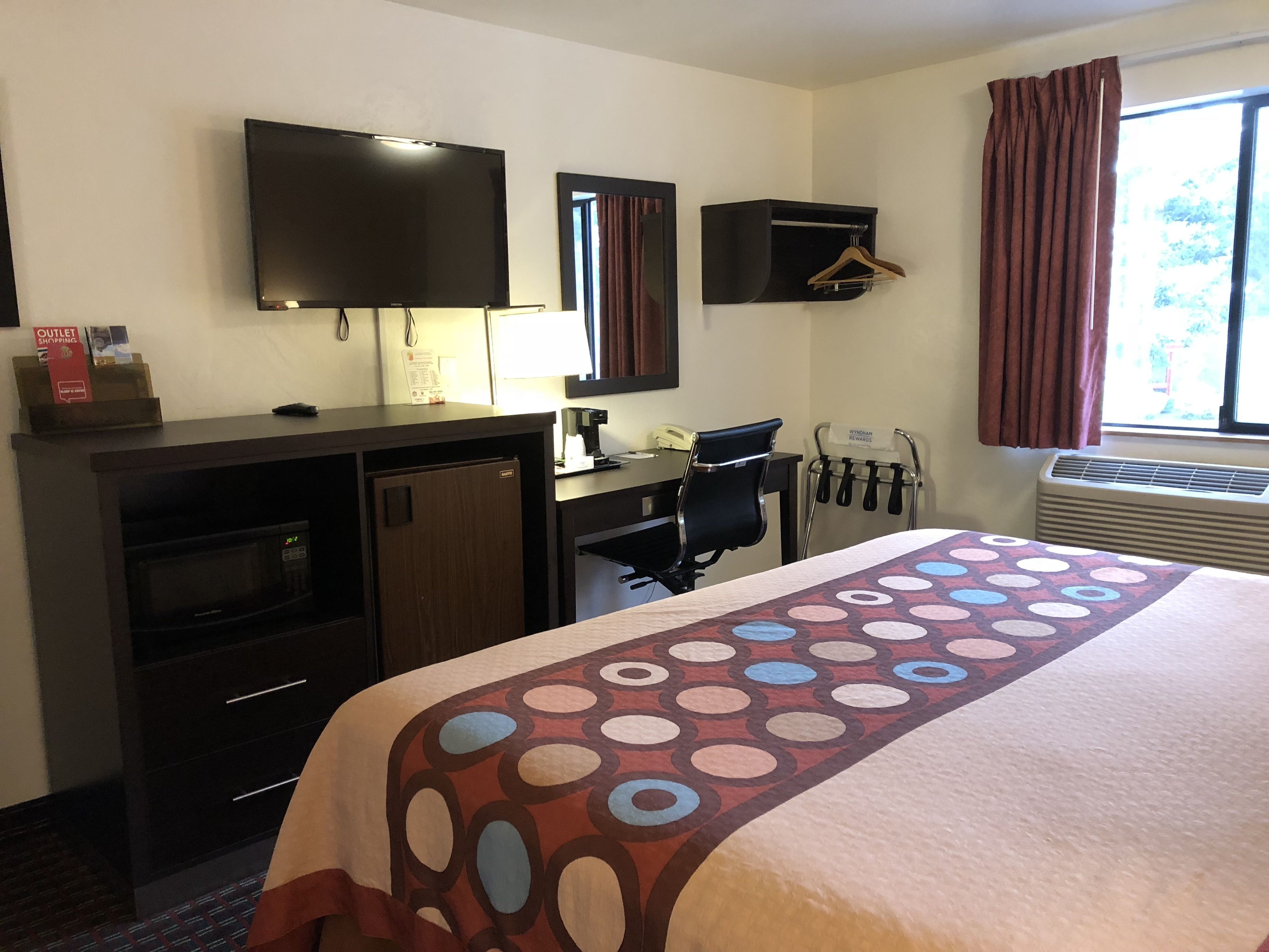 A hotel room with a bed, a desk, a television, a mirror, a coat rack, and an air conditioning unit.
