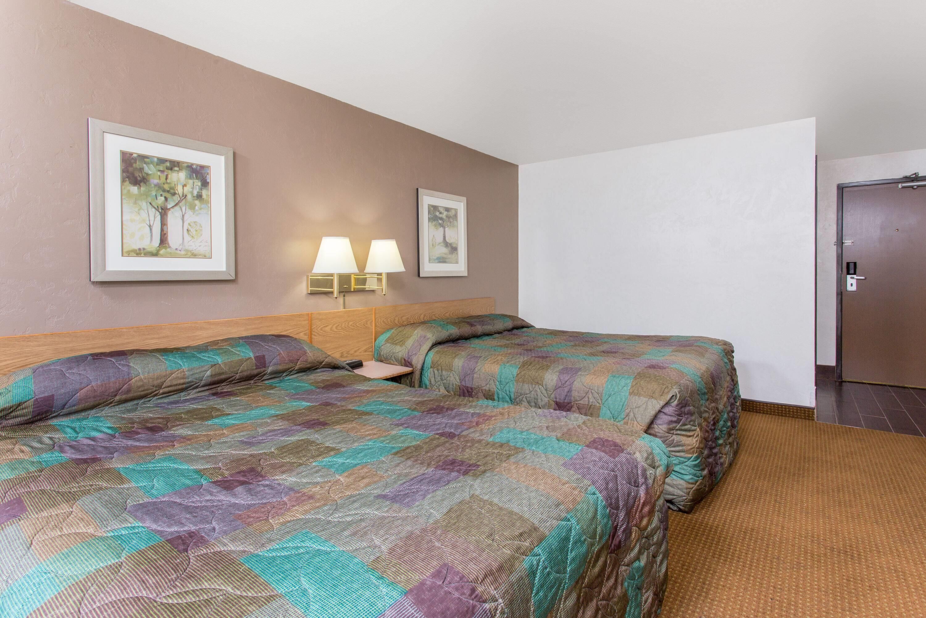 Une chambre d’hôtel avec deux lits, une tête de lit en bois, deux lampes fixées au mur et deux tableaux encadrés accrochés au mur.