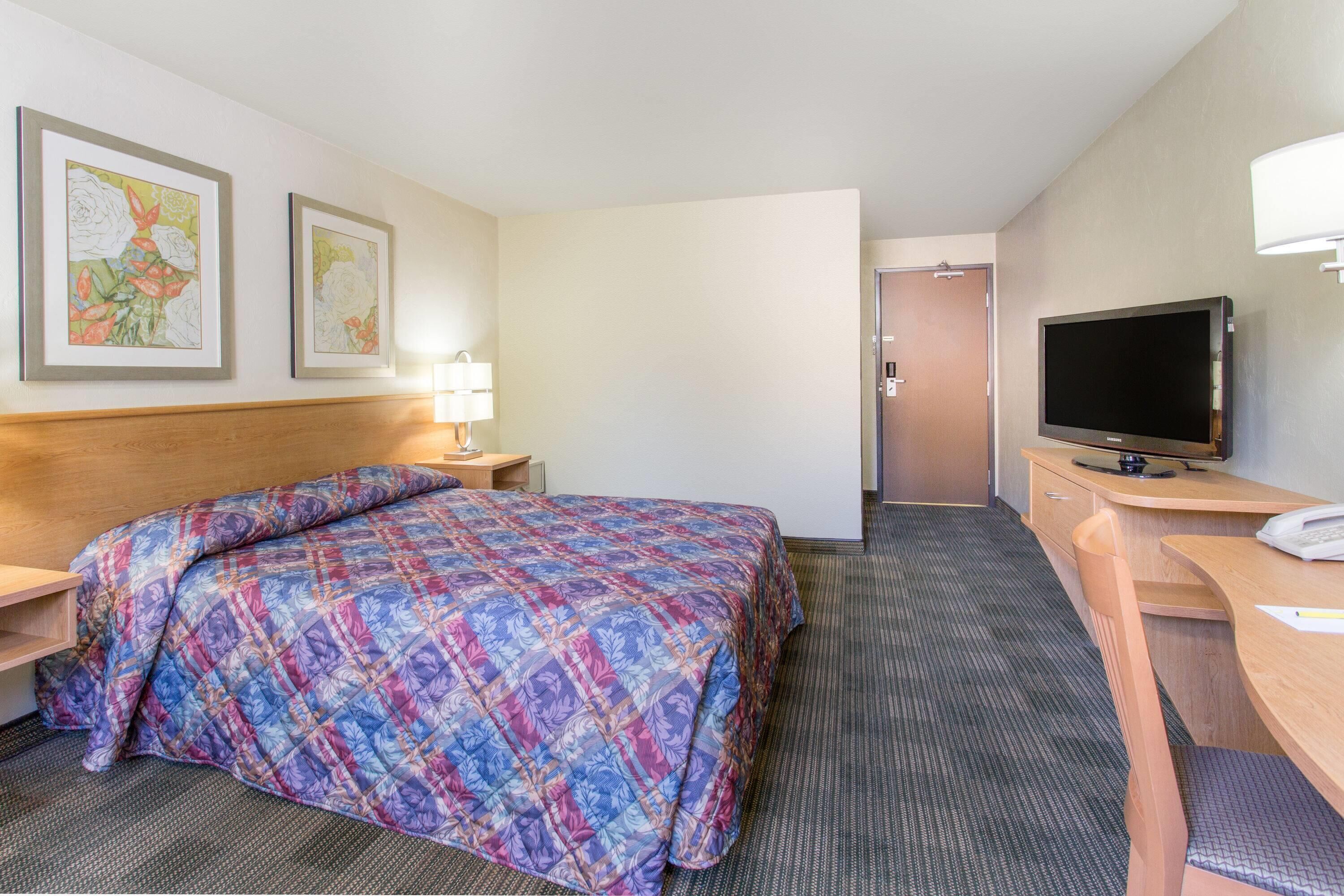 Une chambre d’hôtel comprenant un lit, un bureau, une chaise, une télévision et deux œuvres d’art florales encadrées accrochées au mur.