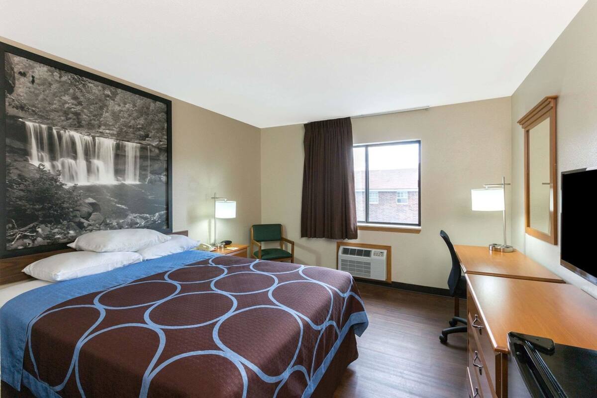 A hotel room with a bed, a desk, a chair, a TV, a window with curtains, and a wall-mounted air conditioner.