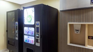 Two vending machines in a room with a wooden panel wall and a small shelf with a triangular sign. - Super 8 by Wyndham Radcliff Ft. Knox Area (Radcliff)