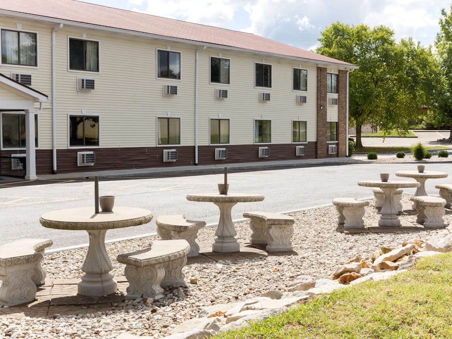 A hotel with a row of outdoor tables and chairs, a covered entrance, and a parking lot.