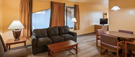 A hotel room with a sofa, a wooden coffee table, a dining table with chairs, a TV, and a lamp.