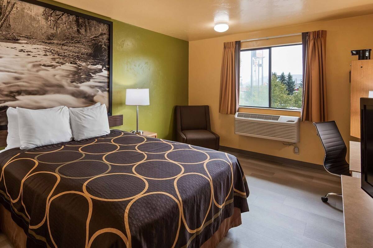 A hotel room with a bed, a desk, a chair, a TV, a window with curtains, and a wall-mounted air conditioner.