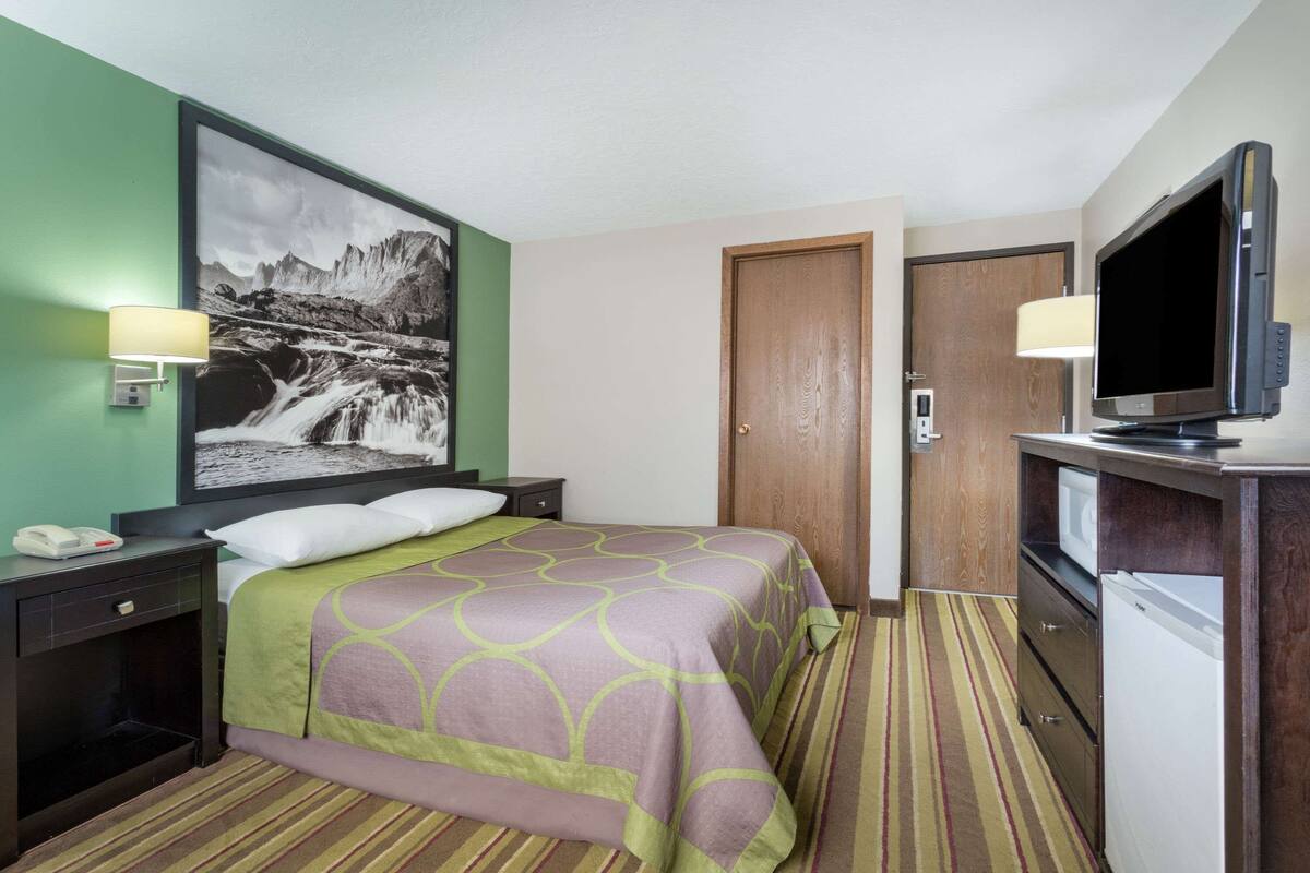 Une chambre d’hôtel équipée d’un lit, d’une télévision, d’un réfrigérateur, d’une table de chevet et d’une lampe fixée au mur.