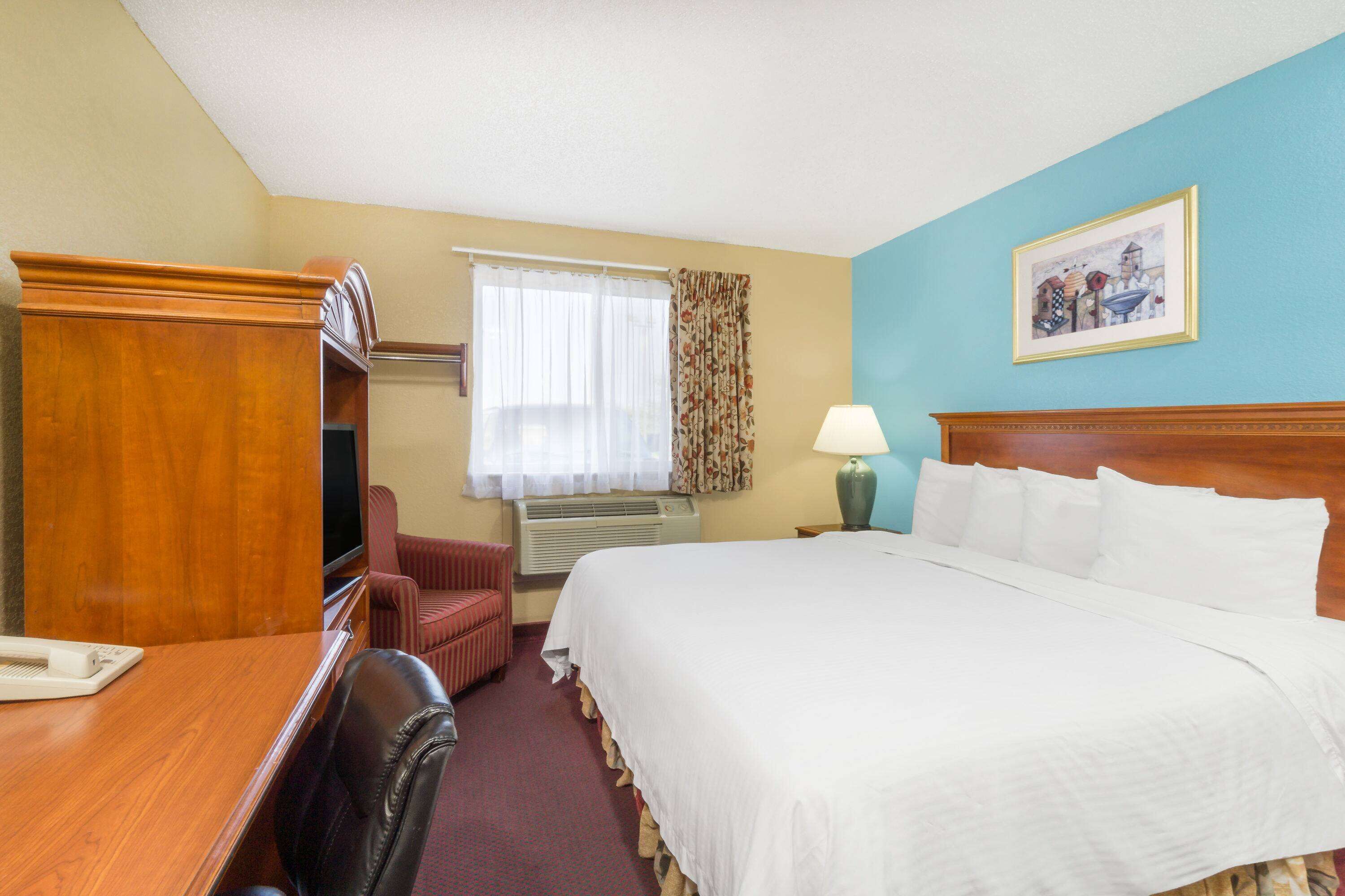 Una habitación de hotel con una cama grande, un escritorio con teléfono, una silla, una ventana con cortinas y un cuadro en la pared.
