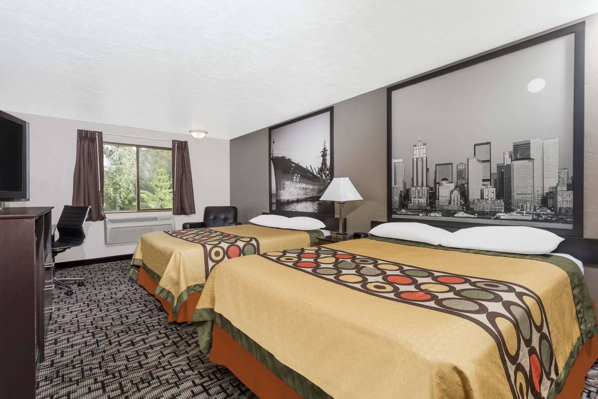 Ein Hotelzimmer mit zwei Betten, einem Schreibtisch mit Stuhl, einem Fernseher, einem Fenster mit Vorhängen und einem Stadtbild-Poster an der Wand.
