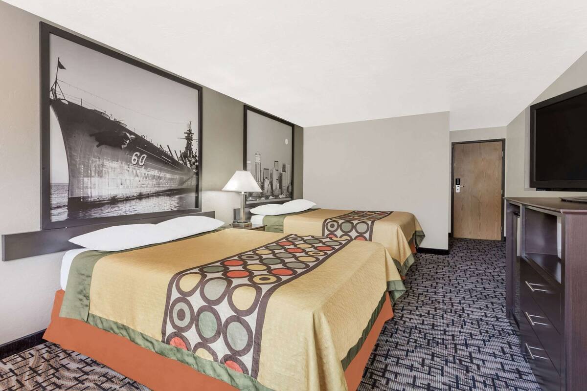 A hotel room with two beds, a TV, a cityscape painting, and a patterned carpet.